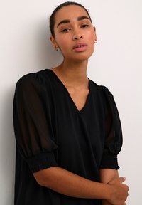 Zwarte blouse met een V-hals en doorschijnende pofmouwen. De mouwen hebben gerimpelde smocking bij de manchetten. Gladde textuur tegen een neutrale achtergrond.