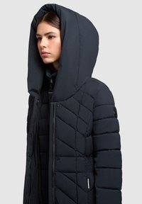 Jonge vrouw draagt een zwart gewatteerd donzen jack met een oversized capuchon, kijkt naar links tegen een effen lichte achtergrond.