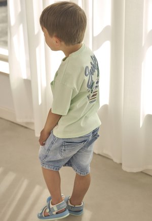 T-shirt in cotone verde chiaro con stampa grafica sul retro, abbinata a pantaloncini di jeans blu e sandali blu. Design minimalista e vestibilità comoda.