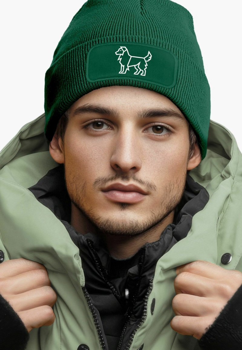Green beanie with ribbed texture and a white embroidered dog design on a rectangular patch; worn with a light green puffer jacket.