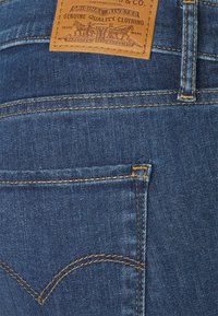 Jeans de mezclilla azul con un parche de marca de cuero color caramelo, remaches de cobre y costuras decorativas amarillas en el bolsillo trasero.
