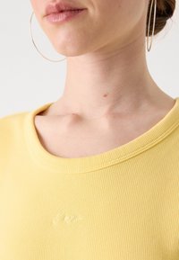 Žluté ribované tričko s kulatým výstřihem a jemným vyšitým logem. Hladká textura s výrazným prošíváním kolem límce.