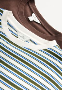 Zwei gefaltete Umstandst-Shirts mit Rundhalsausschnitt; eines einfarbig braun und eines weiß mit grünen und blauen Streifen, die Kleidungsetiketten zeigen.