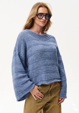 Kobieta ubrana w oversize niebieski dzianinowy sweter, brązowe spodnie i duże kwadratowe okulary przeciwsłoneczne, stojąca z jedną ręką w kieszeni na białym tle.