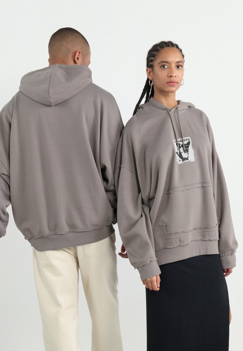 Hoodies cinzentos oversized com bainhas desgastadas, apresentando uma impressão gráfica central. Os modelos estão emparelhados com calças creme e pretas.