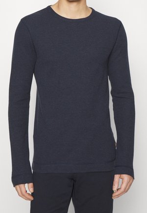 Mand iført ensfarvet marineblå langærmet rundhalset trøje og sorte bukser, stående med armene afslappet langs siden mod hvid baggrund.