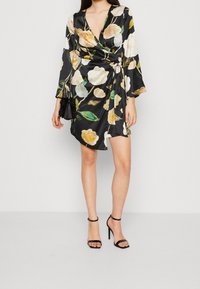 Robe portefeuille florale en satin noir avec des manches longues, ornée de fleurs multicolores et d'un détail de nœud sur le côté. Associée à des escarpins noirs.