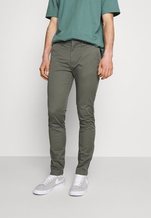 Mężczyzna ubrany w oliwkowe, dopasowane spodnie, morski t-shirt z krótkim rękawem i szare buty Nike, stojący na tle jasnego, jednolitego tła.