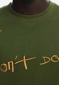 T-shirt in cotone verde khaki con testo ricamato arancione che recita "DON'T DO?" in uno stile scritto a mano. Tessuto morbido, design a collo rotondo.