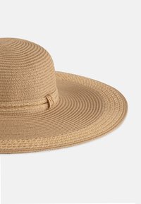 Szeroki kapelusz słomkowy w odcieniu beżowym, z plecioną taśmą. Tekstura jest w formie tkanej, z okrągłymi wzorami na koronie i płaskim rondzie.
