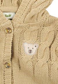 Beige gestrickte Strickjacke mit Kabelmuster, zwei Markenknöpfen und einem kleinen Teddy-Bären-Gesicht-Patch auf der Brust.