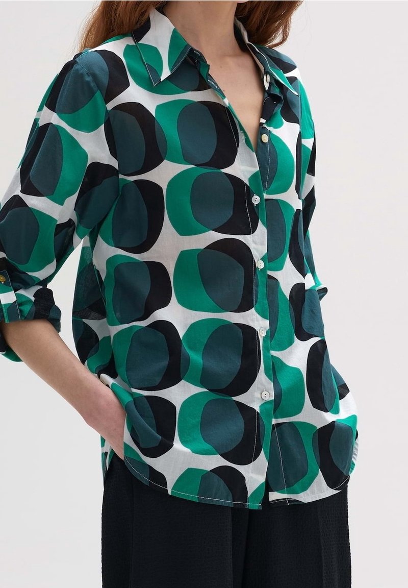 Femme portant une chemise boutonnée avec de grands motifs abstraits de cercles verts et noirs et un pantalon noir, les mains dans les poches, sur fond blanc.