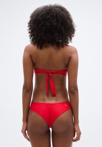 Κόκκινο μπαντό μπικίνι με δέσιμο στην πλάτη και σλιπ χαμηλής μέσης. Υφή υφάσματος, μίνιμαλ σχεδιασμός και διακριτική λεπτομέρεια με λογότυπο στην πλευρά του κάτω μέρους.