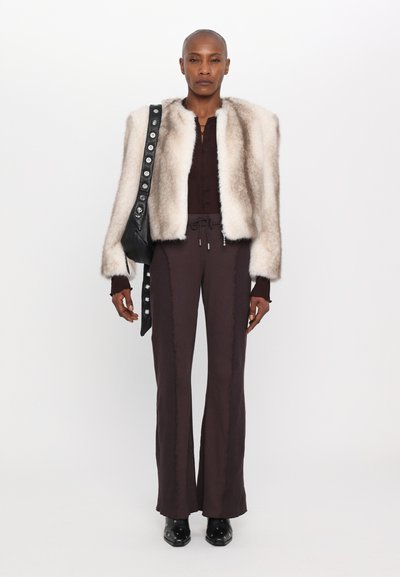 Faux-Fur-Cremewest mit rundem Ausschnitt, kombiniert mit braunen langen Hosen mit seitlichen Nähten. Das Model trägt eine schwarze Tasche mit metallischen Akzenten.