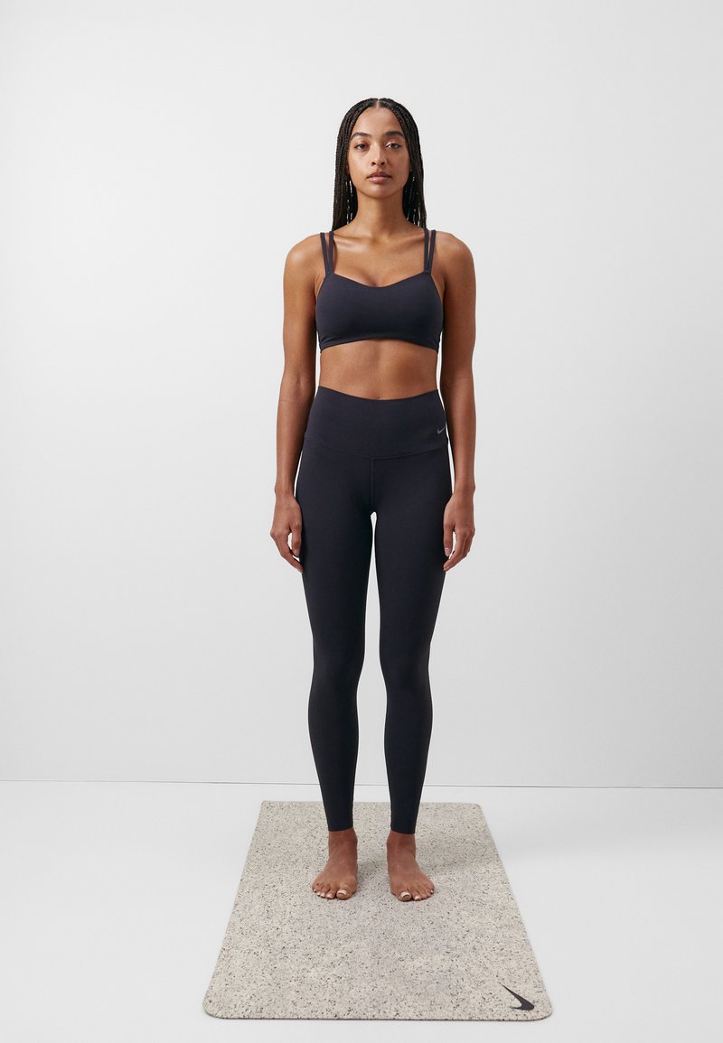 Svart sports BH og høyt liv leggings, begge tettsittende, på en person som står barbeint på et lyst teksturert yogamatte.