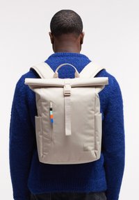 Person wearing a beige roll-top backpack with side pockets and colored zipper pull, dressed in a textured blue sweater, shown from behind.