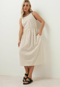 Beige mekko ilman hihoja, jossa on joustava vyötärö, taskut ja mustat yksityiskohdat. Päällä mustat sandaalit, joissa on solkia ja niittejä.