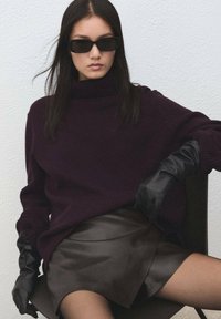 Pulover mov închis cu guler înalt, asortat cu o fustă mini din piele neagră. Modelul poartă ochelari de soare negri și mănuși din piele neagră.