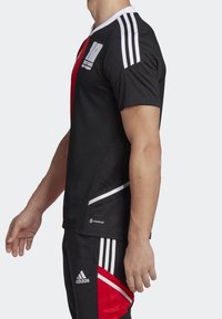 Jersey atlético negro con panel frontal rojo, acentos blancos, tres rayas blancas en los hombros y logo "AEROREADY" cerca del dobladillo.