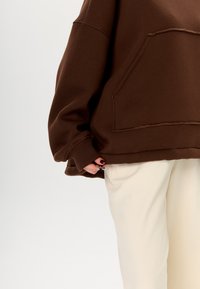 Felpa marrone oversize con tasca frontale, polsini e orlo a coste, indossata sopra pantaloni color crema; texture morbida visibile.