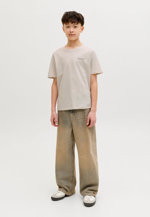 Fiú áll, világos bézs pólót, széles, koptatott barna nadrágot és fehér tornacipőt visel egy egyszerű, világos háttér előtt.