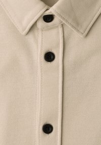 Chemise beige texturée avec un col pointu et trois boutons foncés visibles sur la patte de boutonnage avant.