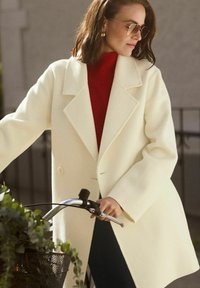 Elfenbensfärgad ullrock med dubbelknapprad, krage och strukturerade axlar, bärs över en röd stickad tröja och matchas med solglasögon.