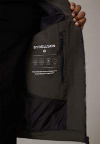 Dunkelolivfarbiger Innenstoff der Jacke mit gedrucktem Markenlogo und Pflegehinweisen; Merkmale umfassen wasserdichtes und winddichtes Material, glatte Textur.