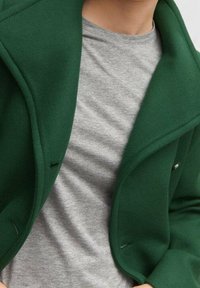 Manteau en laine verte avec une finition texturée et de larges revers, superposé à un t-shirt en coton gris clair. Doté d'un col pointu et de boutons en accents.