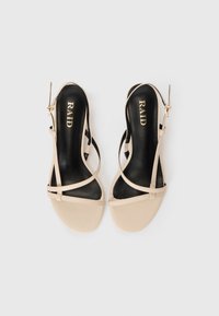 Beige slingback sandaler med sorte indersåler, der har tynde krydsede stropper og en lille hæl. Branding synlig på indersålen.