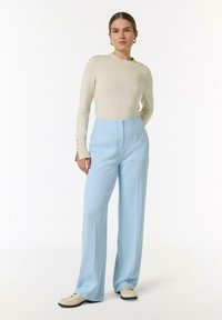Pull en beige clair à côtes avec un col rond et des poignets boutonnés, associé à un pantalon bleu clair taille haute et à jambes larges.