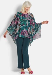 Durchsichtige Blumenbluse in Grün, Pink und Lila mit weit geschnittenen Fledermausärmeln; kombiniert mit dunkeltürkisen Hosen und pinken High Heels Sandalen.