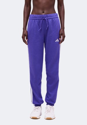 Osoba nosiaca fialové tepláky Adidas s bielymi pruhmi, elastickými členkami a bielymi teniskami, stojaca pred jednoduchým pozadím.