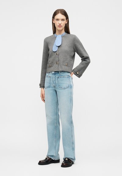 Γυναίκα που φοράει γκρι σακάκι με κουμπιά, μπλε ριγέ κασκόλ, ανοιχτό μπλε ψηλόμεσο τζιν και μαύρα μοκασίνια, στέκεται με το ένα χέρι στη μέση.