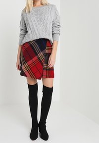 Pull en maille torsadée gris, mini-jupe à carreaux dans des teintes rouges, noires et jaunes, bas noirs au-dessus du genou, textures de tissu lisses.