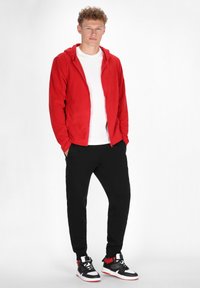 Felpa rossa con zip realizzata in tessuto morbido, abbinata a una t-shirt bianca e pantaloni della tuta neri a gamba affusolata. Presenta scarpe da ginnastica nere e rosse.