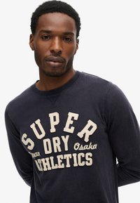 Marineblå sweatshirt med lange ærmer og lys beige "SUPER DRY Osaka ATHLETICS" tekst. Blødt stof med rund hals og broderede detaljer.