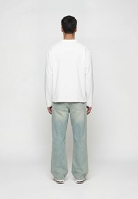 Άνδρας που φοράει λευκό πουκάμισο με μακριά μανίκια, ανοιχτό μπλε τζιν με φαρδιά μπατζάκια και λευκά αθλητικά παπούτσια, στέκεται με την πλάτη γυρισμένη προς το επίπεδο φόντο.