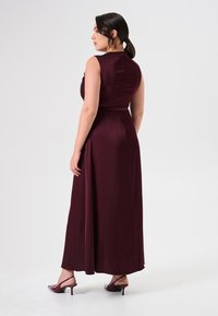 Bordeaux, mouwloze maxi-jurk met een getailleerde bovenkant en een luchtige rok. Gladde stof met een lichte glans. Gecombineerd met doorzichtige hakken.
