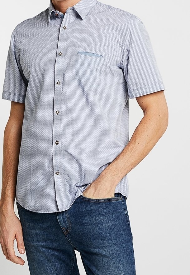 Camisa de manga corta con botones en azul claro, con un patrón texturizado, que incluye un bolsillo en el pecho y botones marrones. Combinada con jeans azules.
