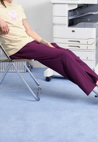 Beige t-shirt med grafiskt tryck, kombinerad med löst sittande lila byxor. Sitter på en kromstol mot en ljusblå matta. Skrivare i bakgrunden.