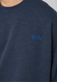 Tmavomodrá mikina vyrobená z mäkkého materiálu, s okrúhlym výstrihom a malým modrým potlačeným logom "American Vintage" na hrudi.