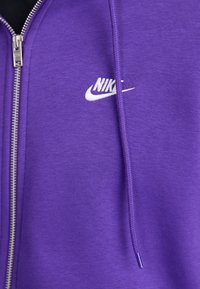 Sweatshirt roxo com fecho éclair, feito de uma mistura de algodão; apresenta um logótipo branco da Nike, fecho éclair prateado e cordão ajustável para o capuz.