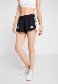 Kvinna klädd i svarta Adidas träningsshorts, vit sport-bh och vita sneakers, stående mot en enkel ljus bakgrund.