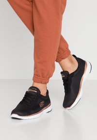 Czarne sportowe buty z akcentami w kolorze różowego złota, siateczkowa cholewka zapewniająca oddychalność oraz biała podeszwa, zestawione z pomarańczowymi dresami ze ściągaczami.