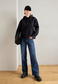 Black cropped hoodie with a front pocket, paired with loose blue denim jeans and black platform shoes. Holding a black pleated bag.