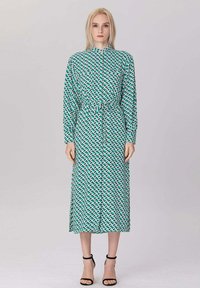 Une femme blonde se tient debout portant une robe midi à manches longues, verte et blanche avec un col montant et des talons à bride de cheville noirs.