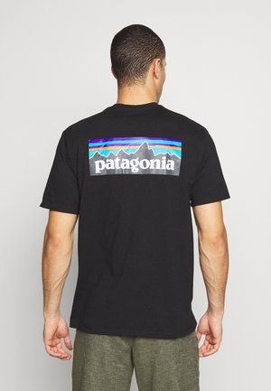 Homme de dos portant un t-shirt noir Patagonia avec un logo de montagne coloré et un short gris-vert sur un fond uni.