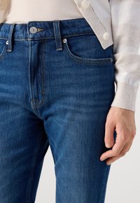 Nærbilde av en person med mørkeblå jeans og en lys kardigan, der den ene hånden hviler på låret.