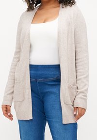 Béžový pletený kardigan s otevřeným předním dílem, žebrovanými manžetami a dvěma předními kapsami, v kombinaci s modrými džínami s vysokým pasem a bílým topem na ramínka.